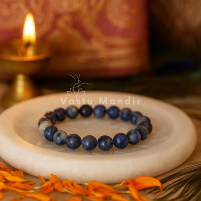 100% Natural Sodalite beads with deep blue color and white veining on a white plate with 'Vastu Mandir' branding, candle in the background