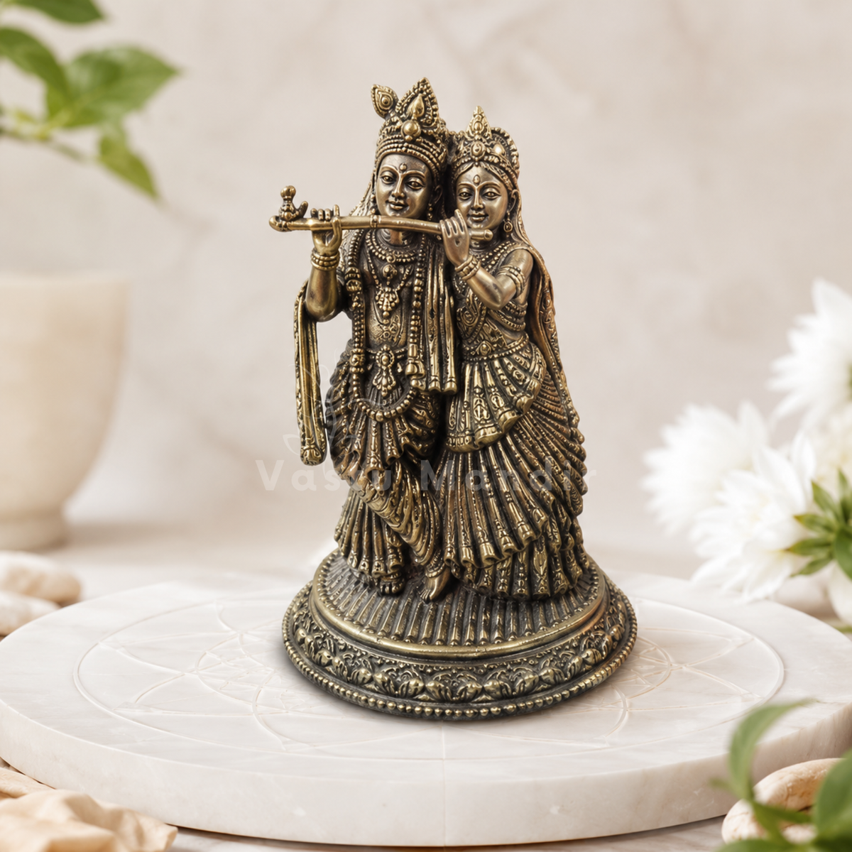 Brass Radha Krishna idol displayed on a white pedestal beside flowers