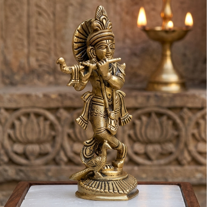 Brass Krishna Ji idol displayed in a carved temple-style setting beside a diya