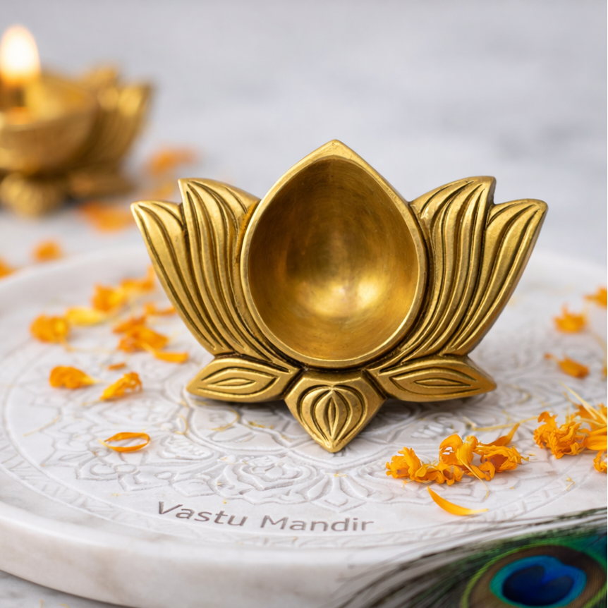 Gold lotus-shaped candle holder on a marble base with 'Vastu Mandir' branding, surrounded by orange flowers and a peacock feather.