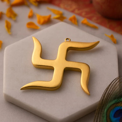 A gold swastika pendant rests on a beige stone surface adorned with orange petals and a peacock feather.