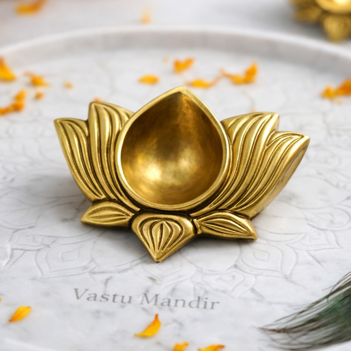 Brass lotus-shaped diya on a marble plate with marigold flowers and a peacock feather.