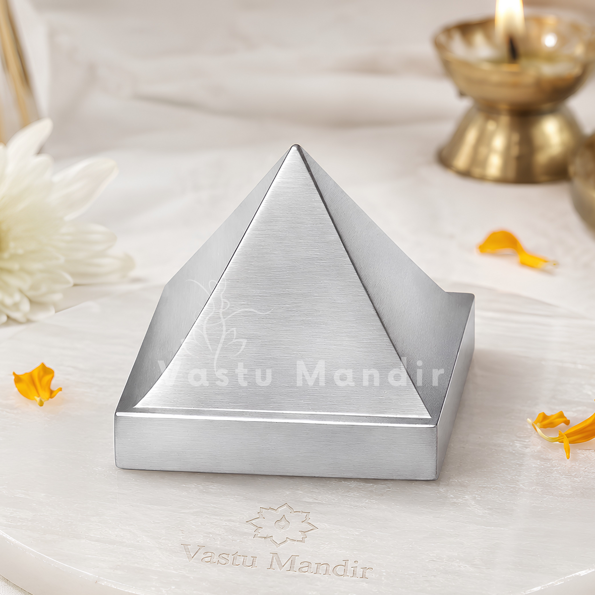 An aluminium pyramid placed on a round white marble tray, styled with a lit brass oil lamp, a white flower, and scattered orange petals in a soft indoor setting.