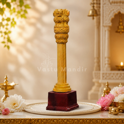 A Wooden Ashoka pillar statue on a dark red wooden base, displayed on a white plate with decorative flowers and traditional lamps in a warm, ornate setting.