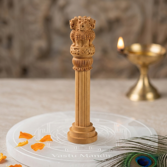 Wooden Finish Ashoka Pillar Idol Vastu Remedy on a white base with 'Vastu Mandir' branding, surrounded by orange petals and a peacock feather.