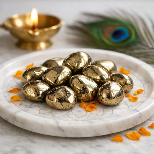 Golden pyrite tumbled stones arranged on a carved white plate with marigold petals and a softly lit diya