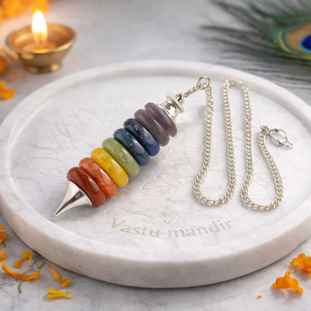 Seven chakra pendulum with silver chain on a white marble plate beside a peacock feather