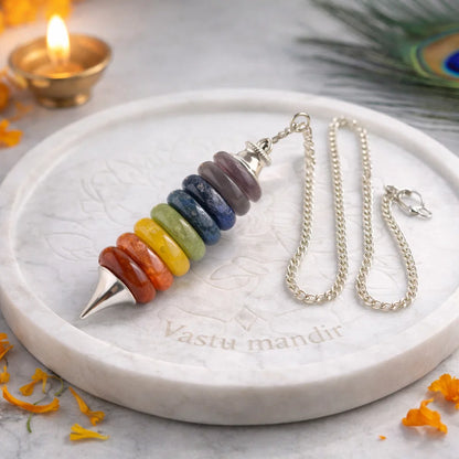 Seven chakra pendulum with silver chain on a white marble plate beside a peacock feather